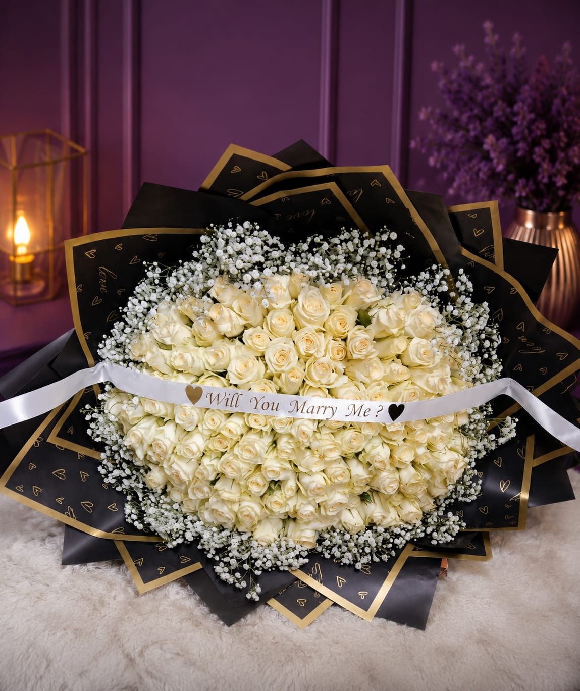 Luxury white rose proposal bouquet with Will You Marry Me sash in black and gold heart-print wrap with baby's breath on white fur rug against purple wall