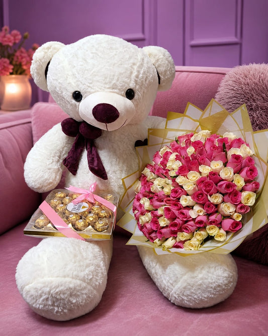 Giant white teddy bear with burgundy bow holding Ferrero Rocher chocolates next to 100 pink and white roses bouquet in gold wrap on pink velvet sofa